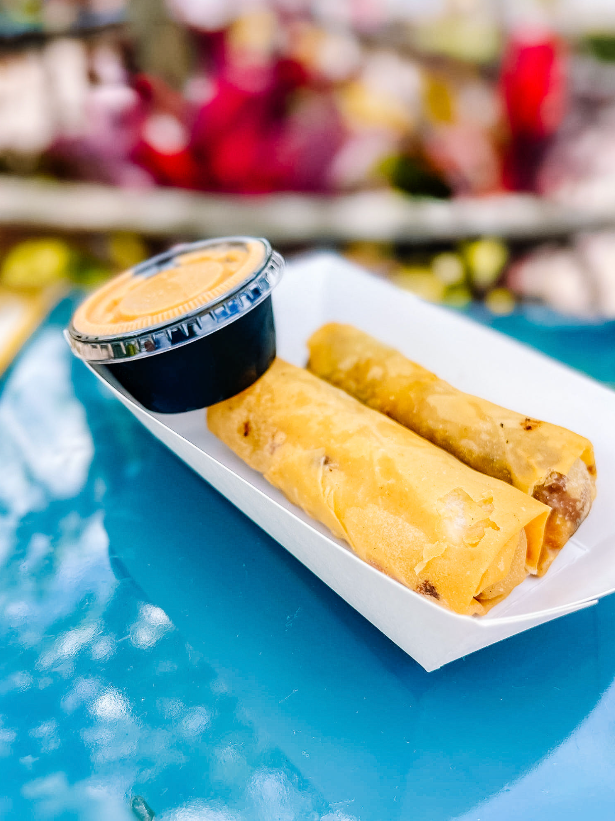 Spring rolls served on a white paper tray with a cup of dipping sauce on a turquoise surface.