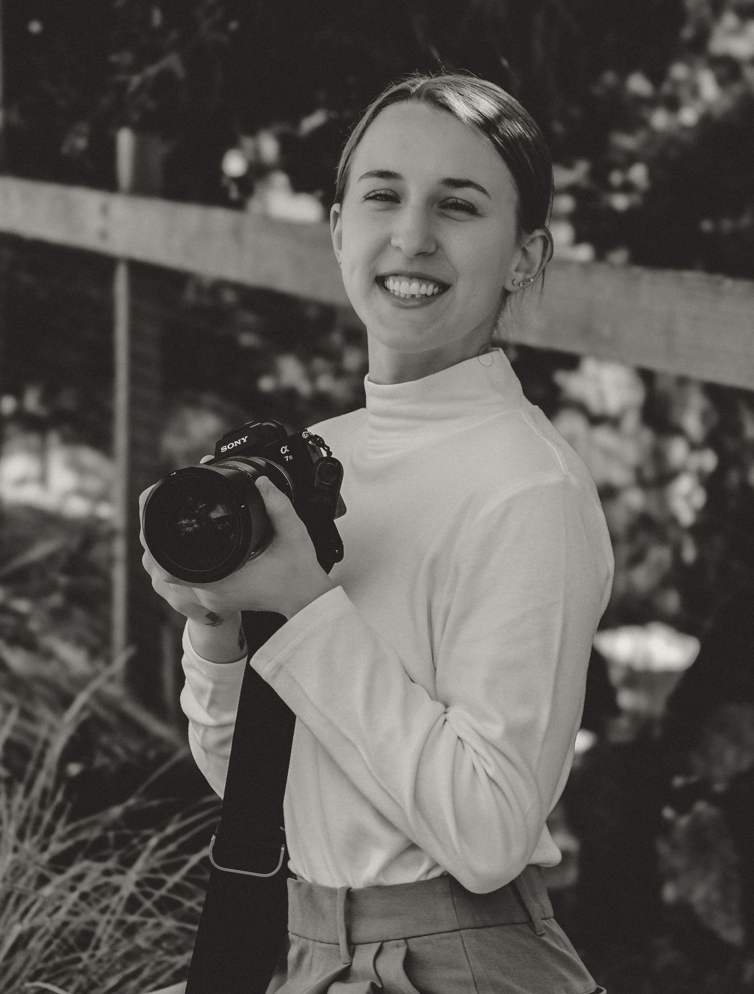 A photographer in a white turtleneck sweater smiles while holding a camera outdoors in black and white.