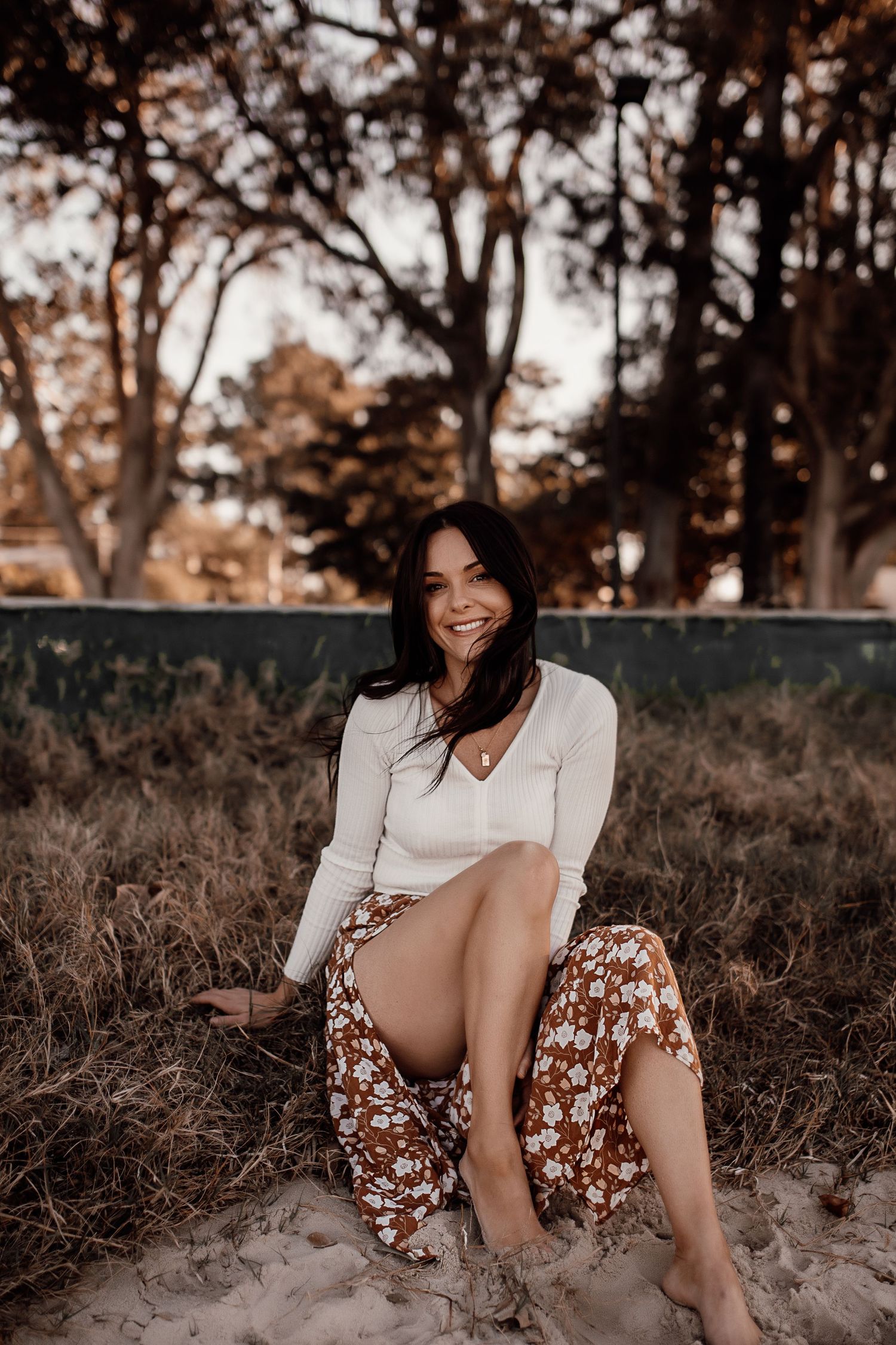Outdoor portrait with warm autumn colors showing someone in white sweater and floral skirt sitting on sandy ground.