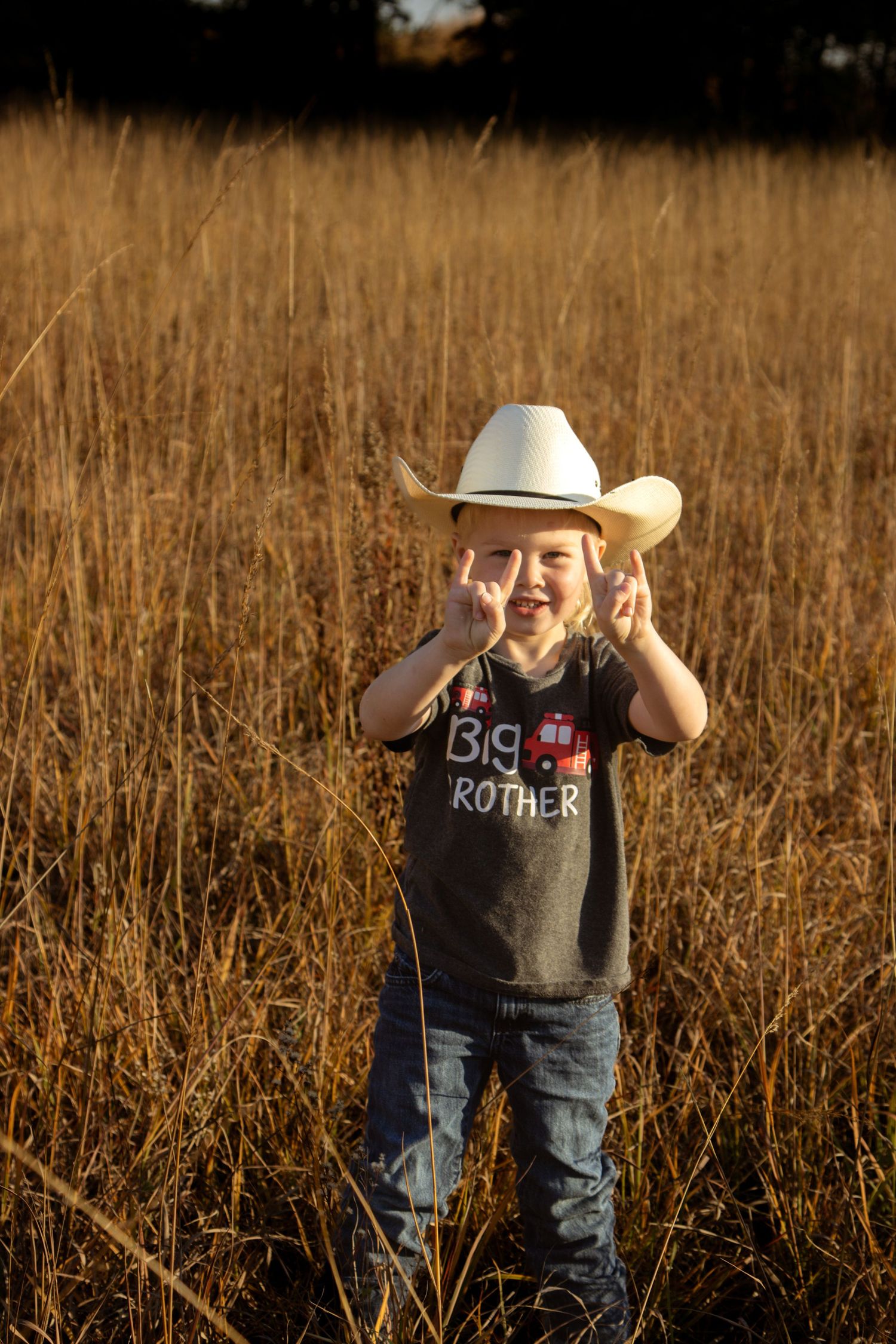 A young child in a cowboy hat and jeans stands in a golden field at sunset.
