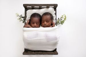 Two sleeping babies tucked in white blanket on brown wooden bed with green floral decorations.