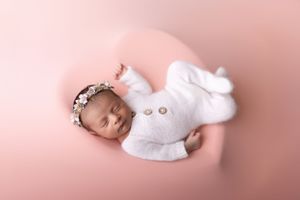 Sleeping newborn baby wearing white knit outfit and floral headband on soft pink background.