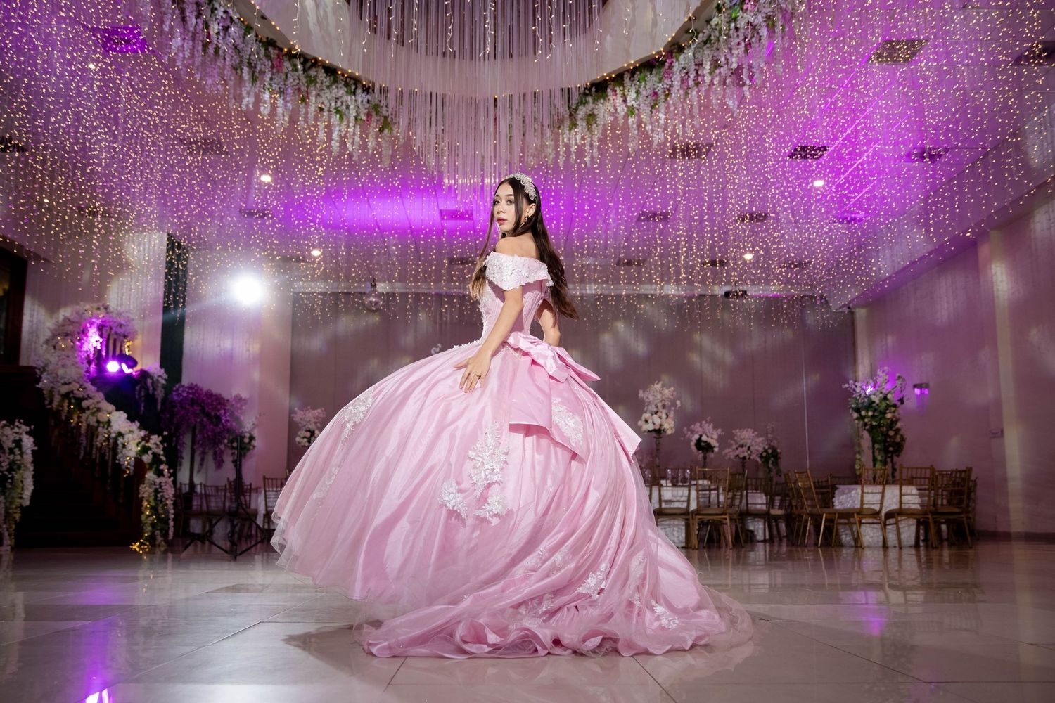 Elegant pink ballgown illuminated by purple lights in a grand ballroom with crystal chandeliers.