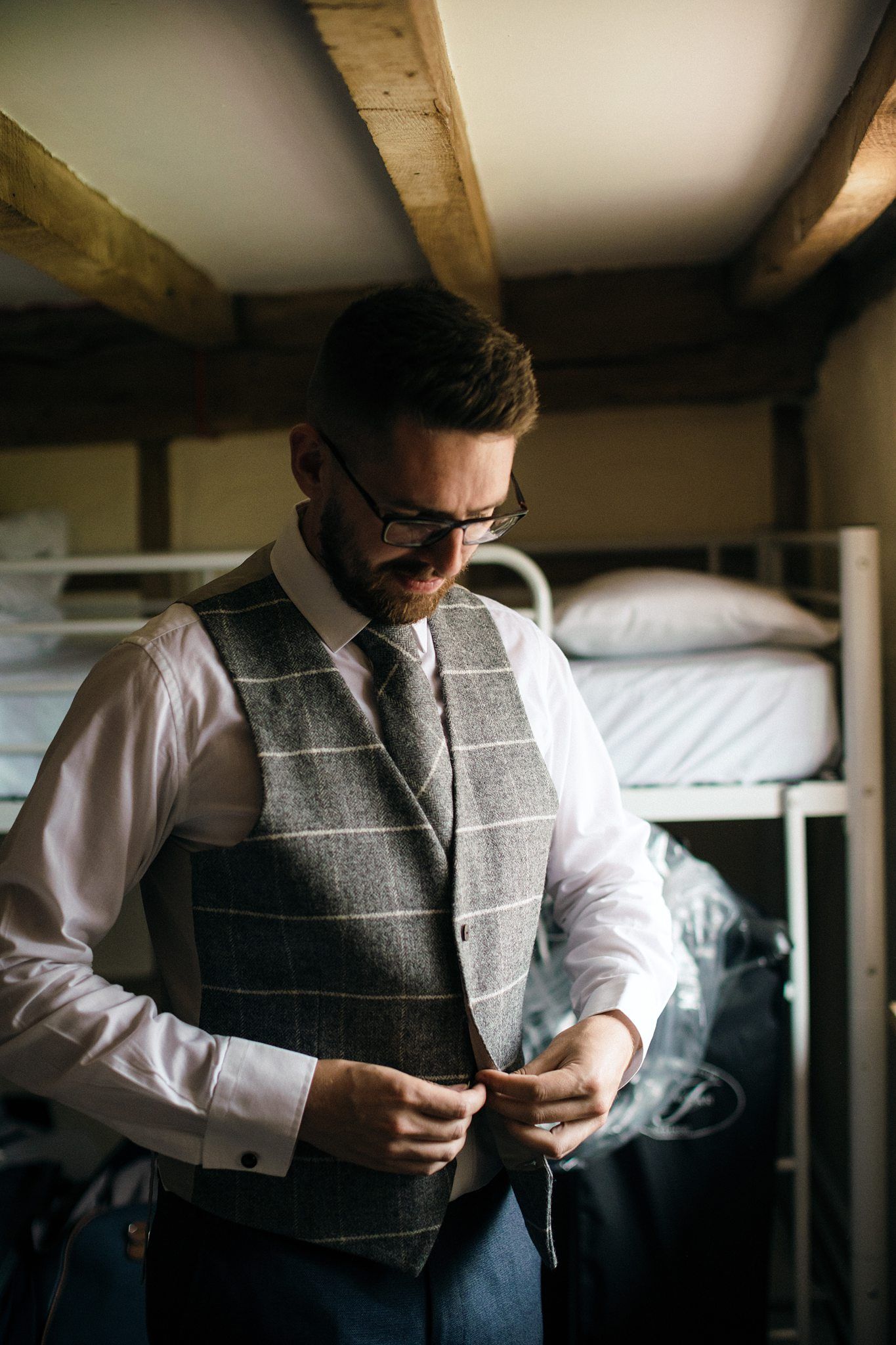 A formal attire scene shows a groom adjusting details while wearing a plaid vest and dress shirt in a rustic room at Healy Events, Kentville