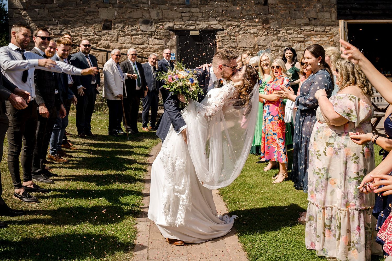 Wedding guests shower confetti on a celebratory couple kissing on a sunny day in front of Healy Events building, Kentville Nova Scotia