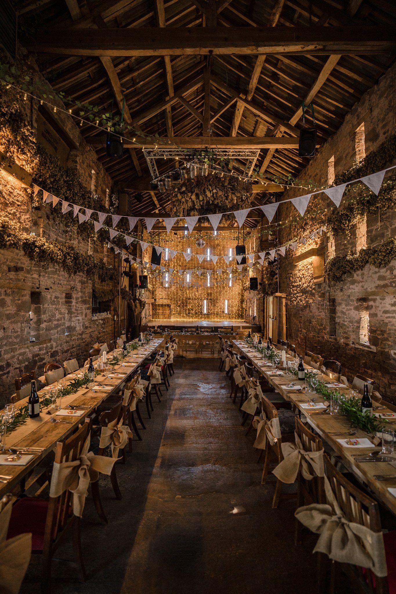 A rustic Healy Events stone barn decorated for a wedding reception with string lights and bunting flags above long wooden dining tables.