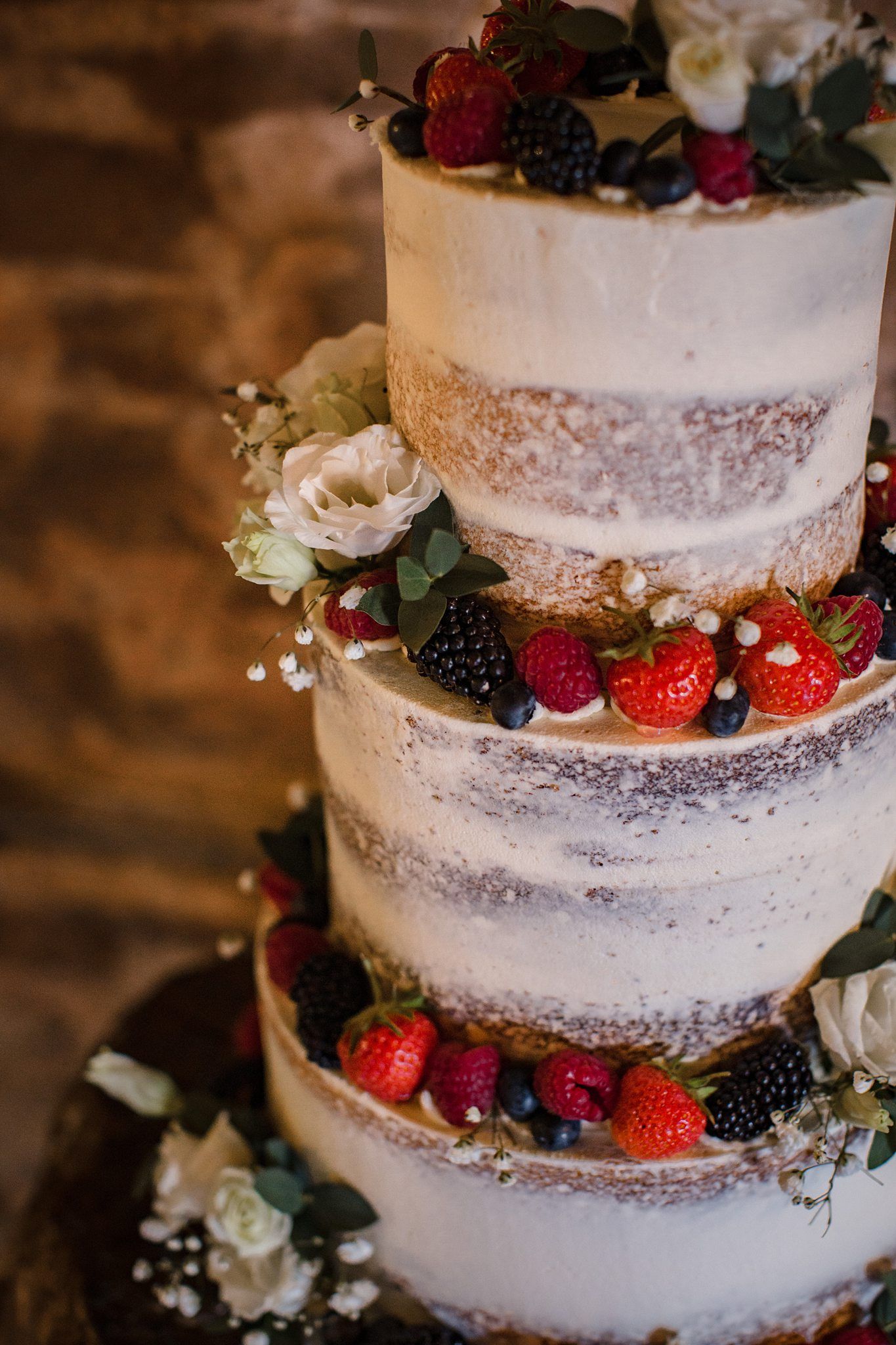 A rustic three-tiered semi-naked wedding cake decorated with fresh berries, roses, and eucalyptus leaves in the barn at Healy Events