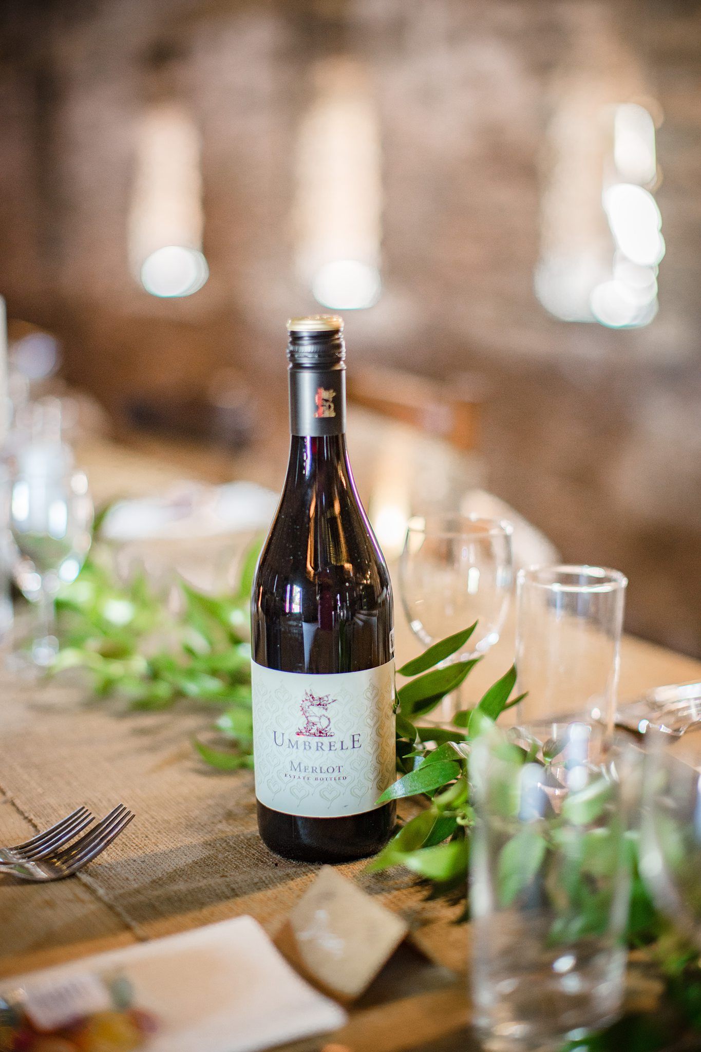 Red wine bottle on rustic wooden table at Healy Events wedding venue with greenery and glassware at elegant dining setup with bokeh lights.