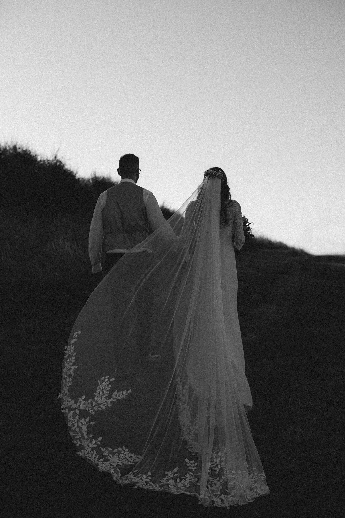 A black and white silhouette of a bride in a flowing lace veil and groom walking up a hillside at dusk near Healy Events. in Kentville, NS.