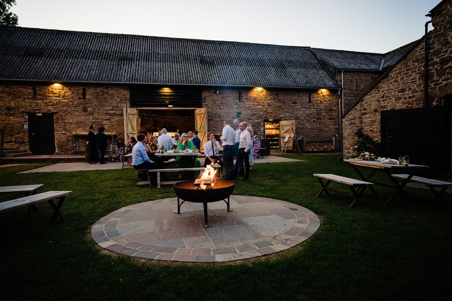 People gather around a fire pit on a circular stone patio at dusk outside a rustic barn with warm lighting at Healy Events wedding venue.