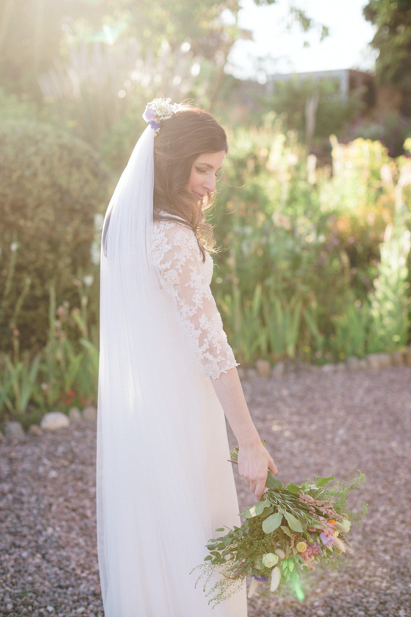 Bride in lace gown with long veil holding bouquet in garden setting with sunlight at Healy Events wedding venue gardens.
