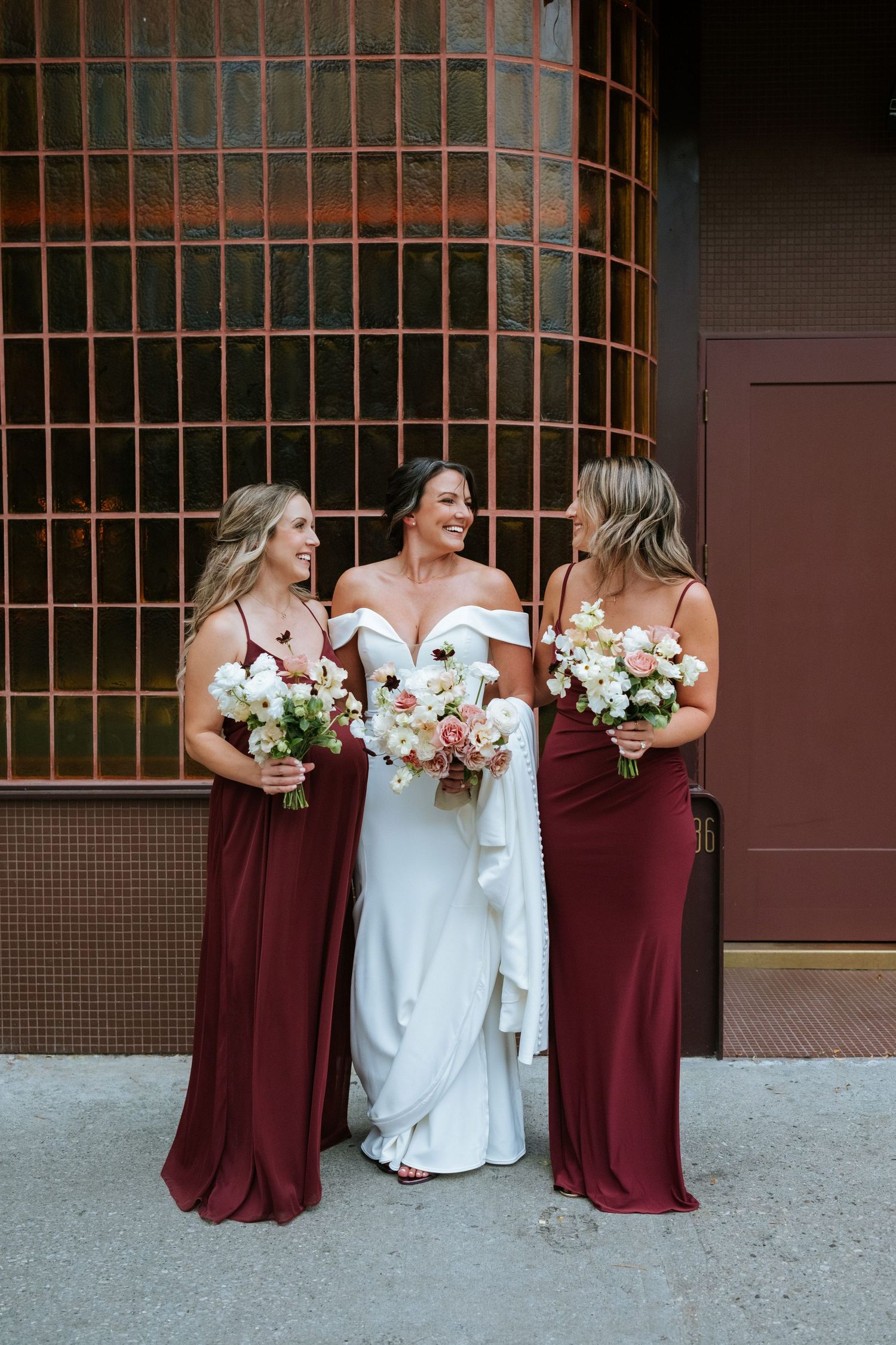 Bridesmaids in burgundy dresses hold bouquets while posing with bride against brick wall.