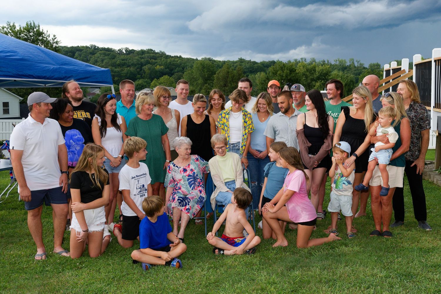 Large multi-generational group gathered outdoors on grassy area during summer birthday event.