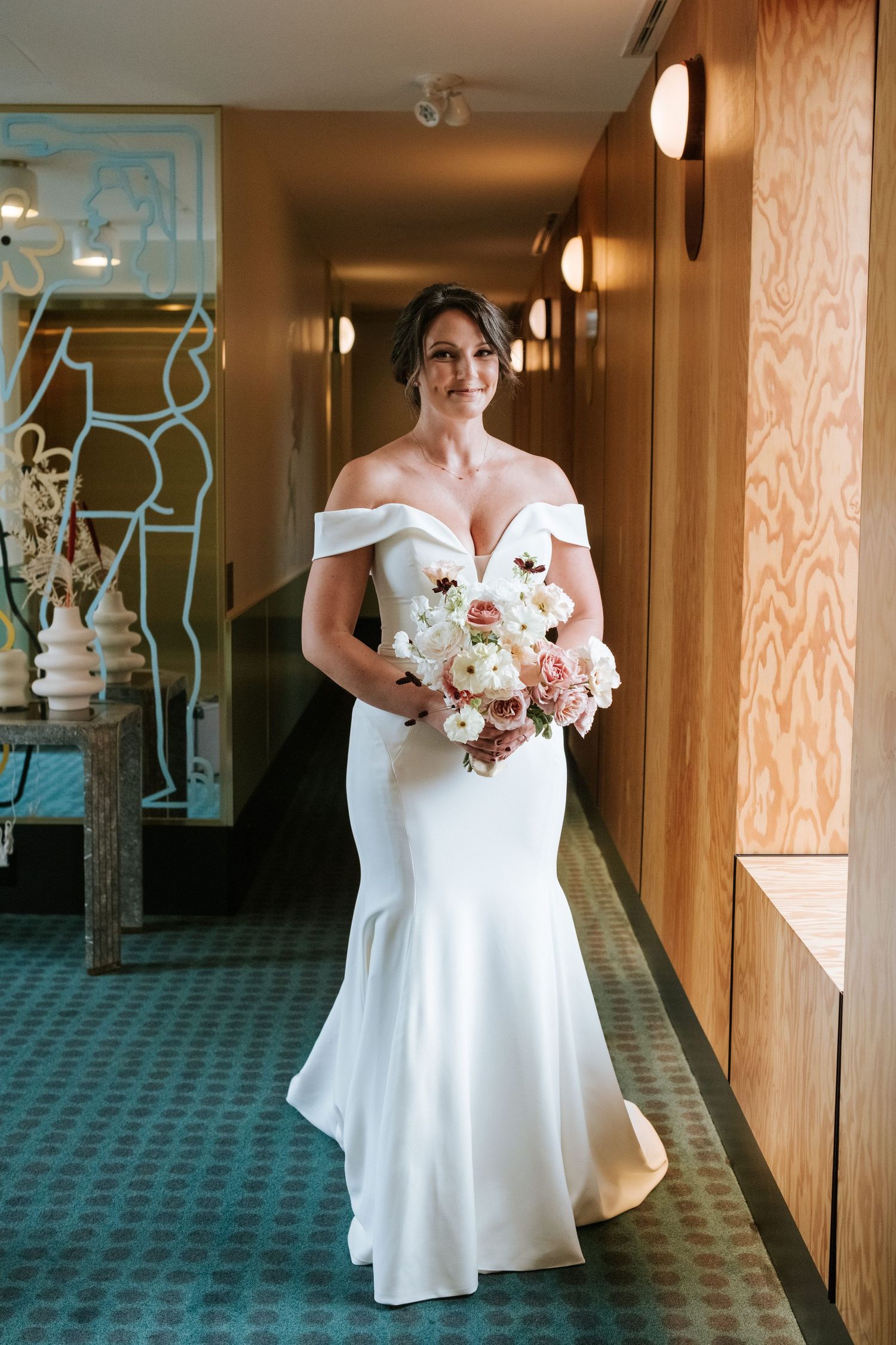 Elegant bride in white off-shoulder mermaid gown holding pink bouquet in hotel hallway.