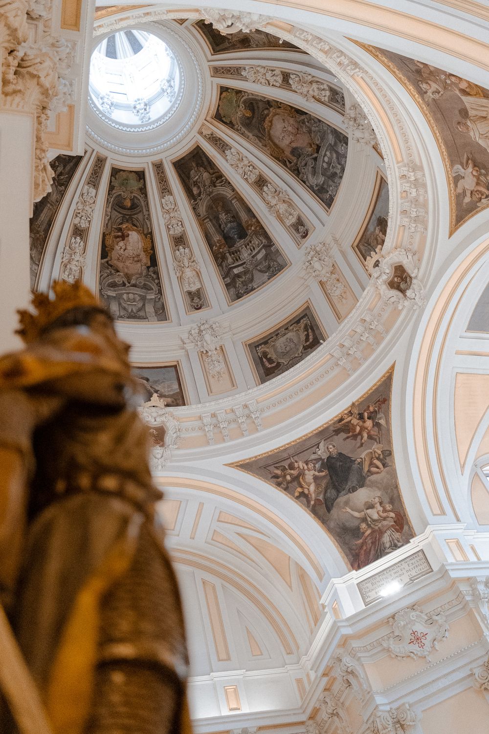 Ornate baroque church dome interior with detailed white plasterwork and golden statue detail.