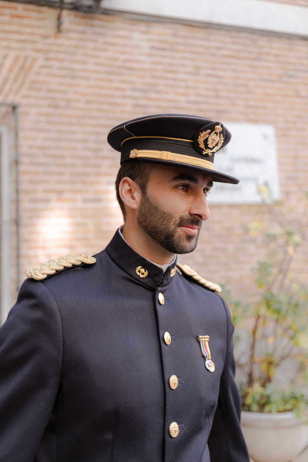 Naval officer in formal dress uniform with cap and medals against brick wall.