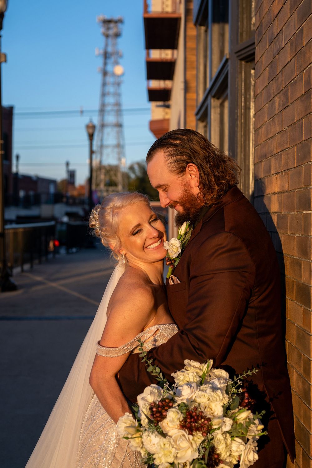 A couple shares an intimate moment together at sunset against a brick building with a beautiful floral bouquet.