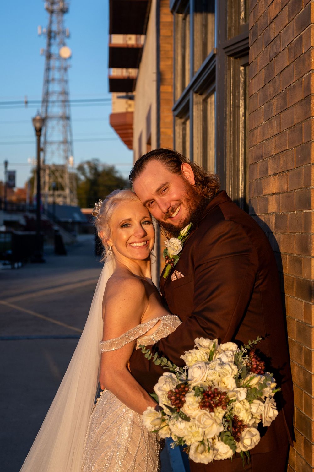 A bride and groom share a romantic moment against a brick wall during golden hour light, with a floral bouquet of white blooms.