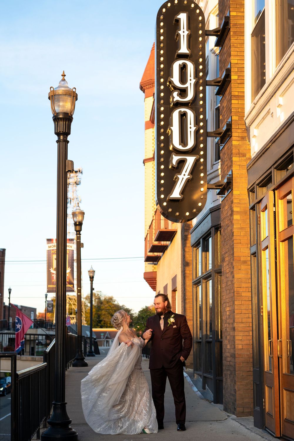 A couple stands together under a vintage 1907 sign at dusk, with decorative street lamps illuminating the historic building.