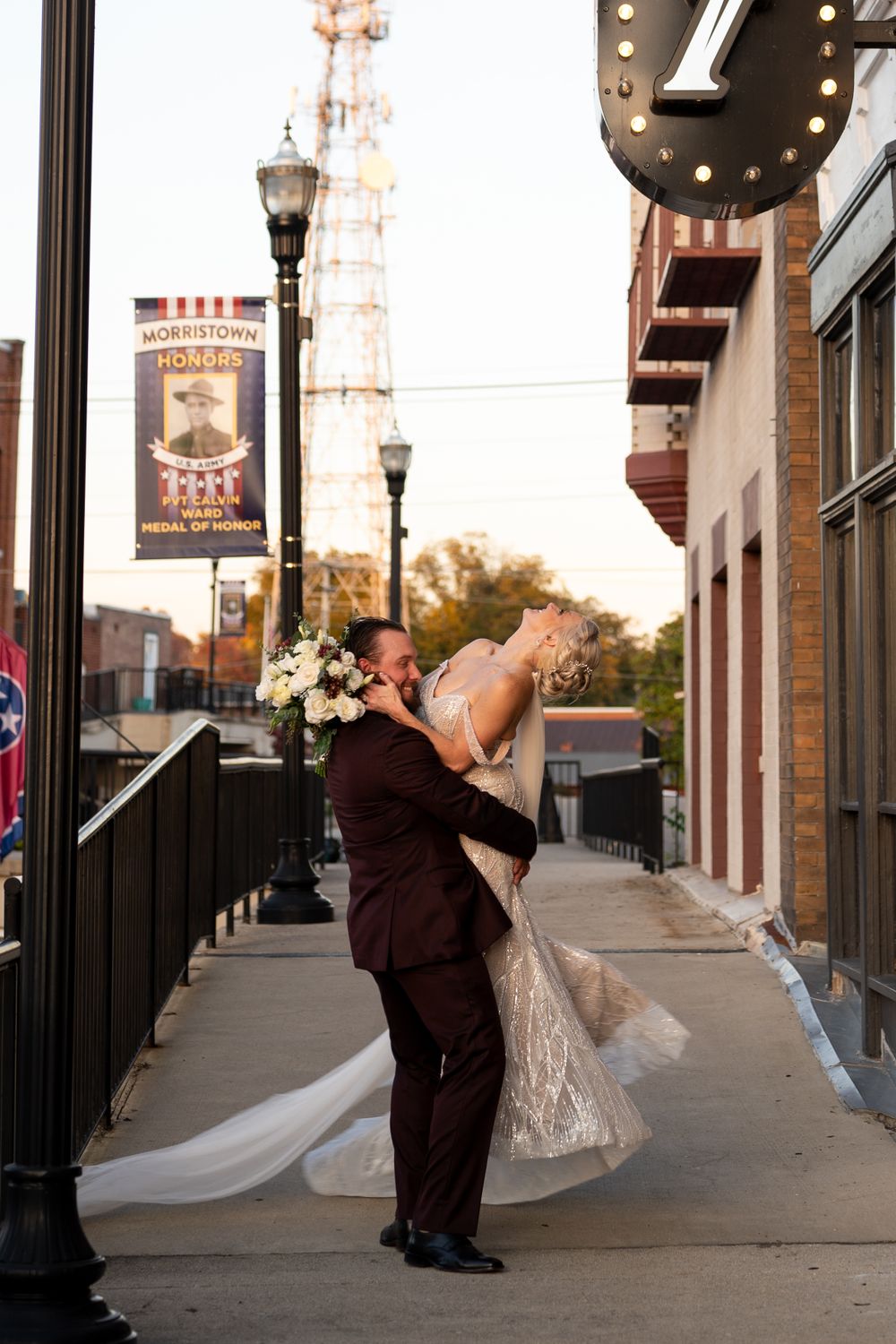 A romantic moment between a couple in formal attire on a city sidewalk at sunset with vintage lampposts and storefronts.