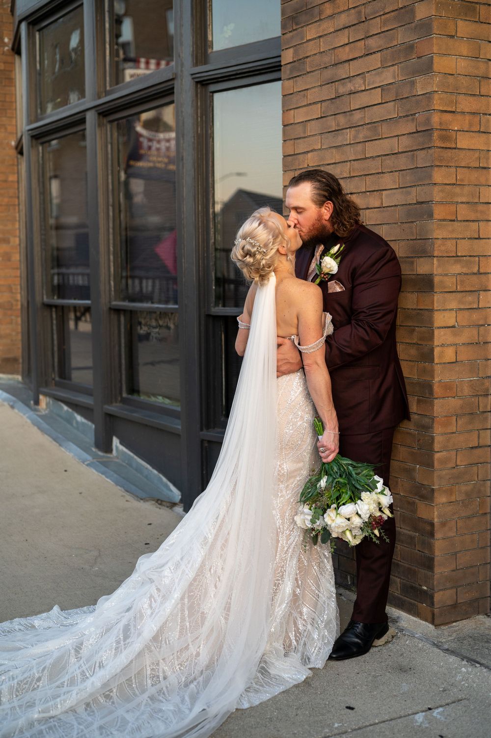 A romantic wedding moment against a brick wall with a white lace gown and burgundy suit.