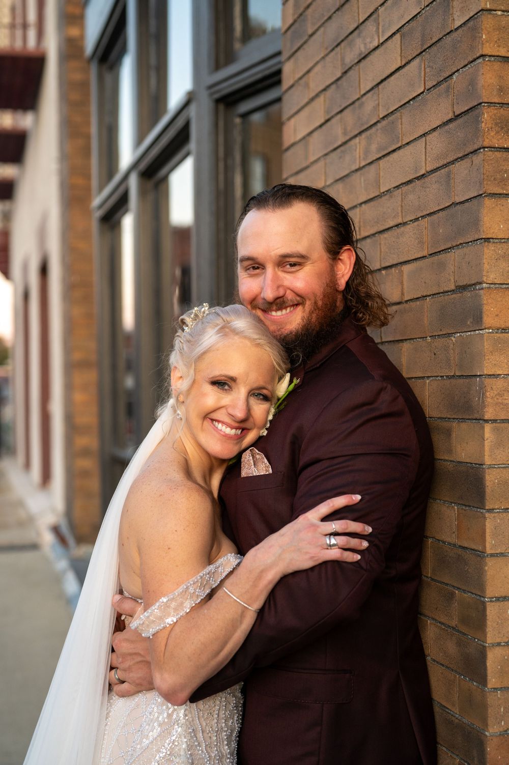 A happy bride and groom embrace against a brick wall on their wedding day, sharing a tender moment together.