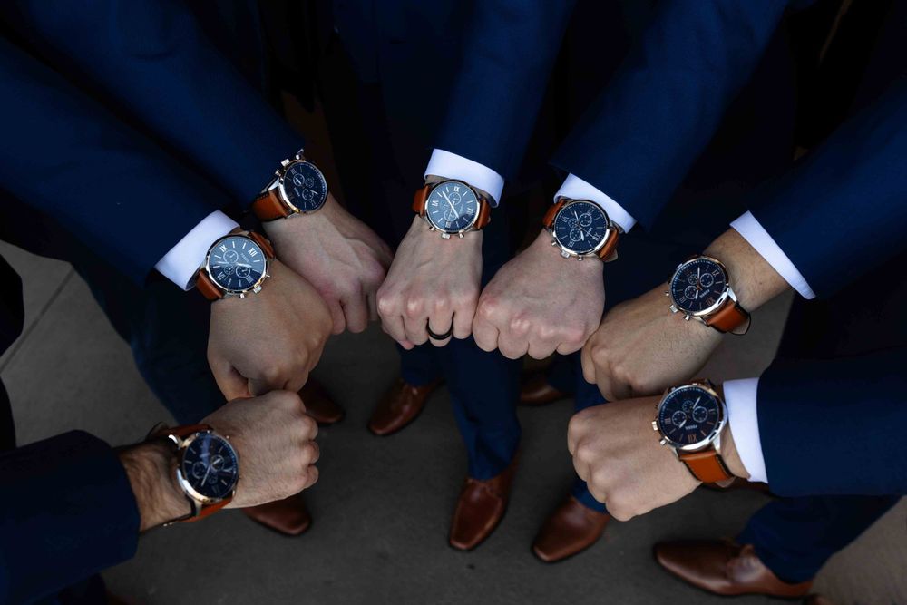 Groomsmen in navy suits join hands in a circle showing their matching black watches against white shirt cuffs.