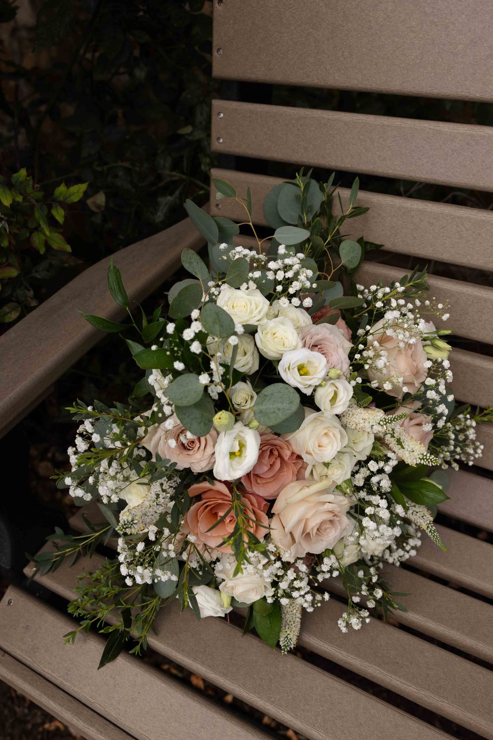 A romantic wedding bouquet with blush pink and white roses, baby's breath, and eucalyptus leaves rests on a wooden bench.