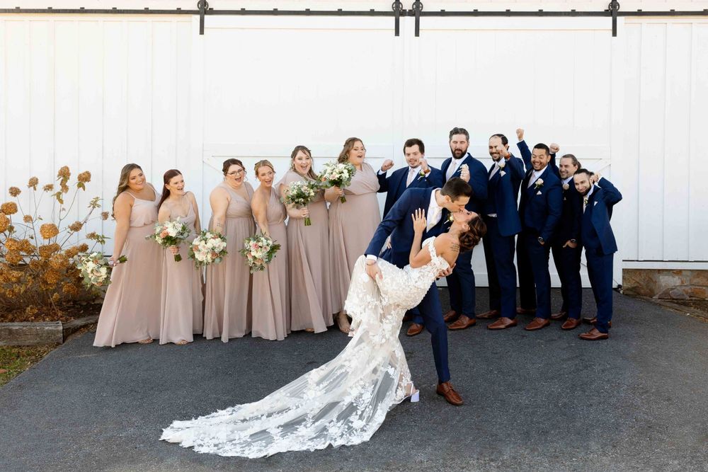 Wedding party poses playfully against white barn wall with bridesmaids in beige and groomsmen in navy blue suits.