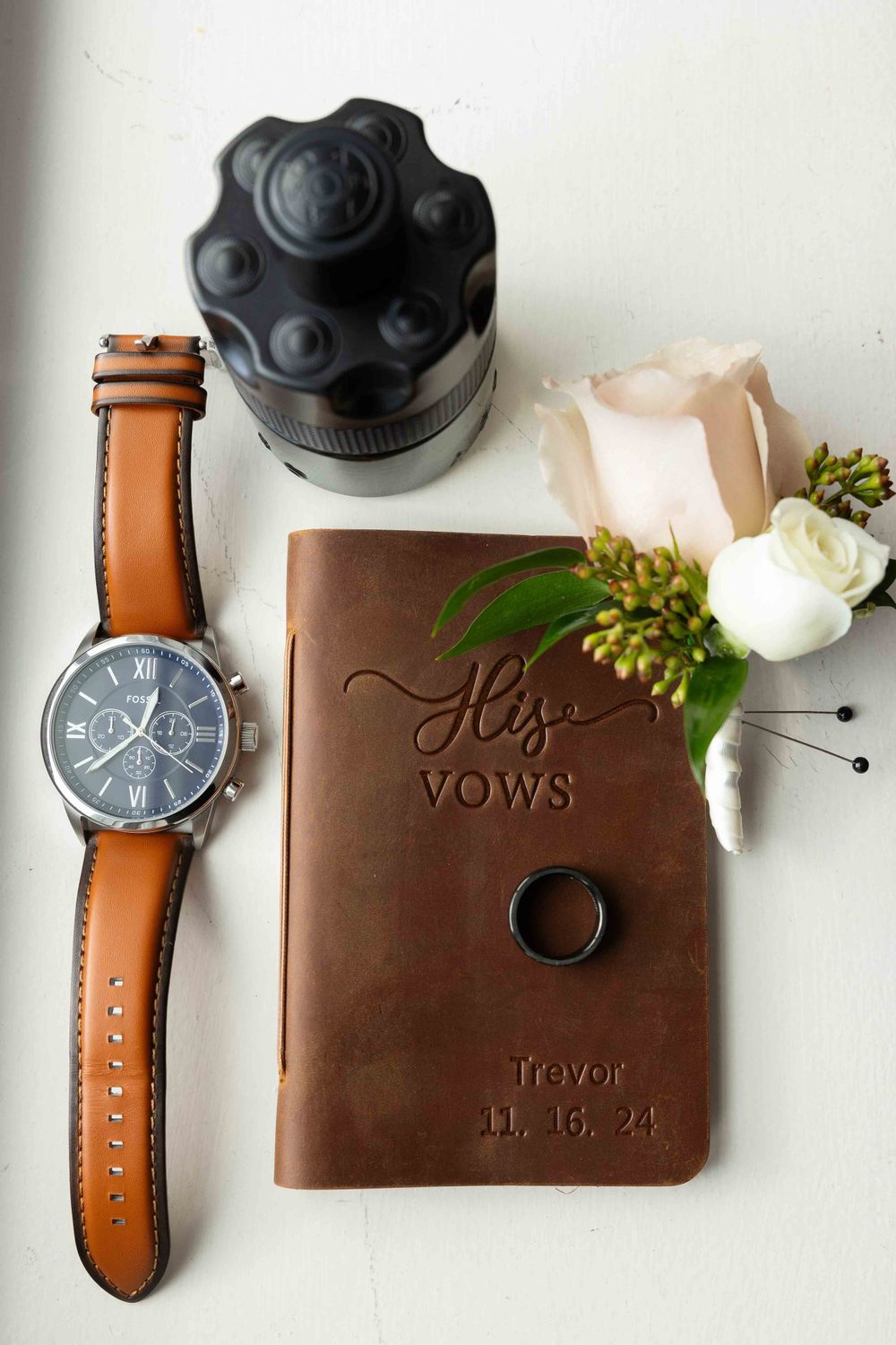 Wedding vows book with brown leather cover displayed alongside watch, flowers and camera lens on white surface.