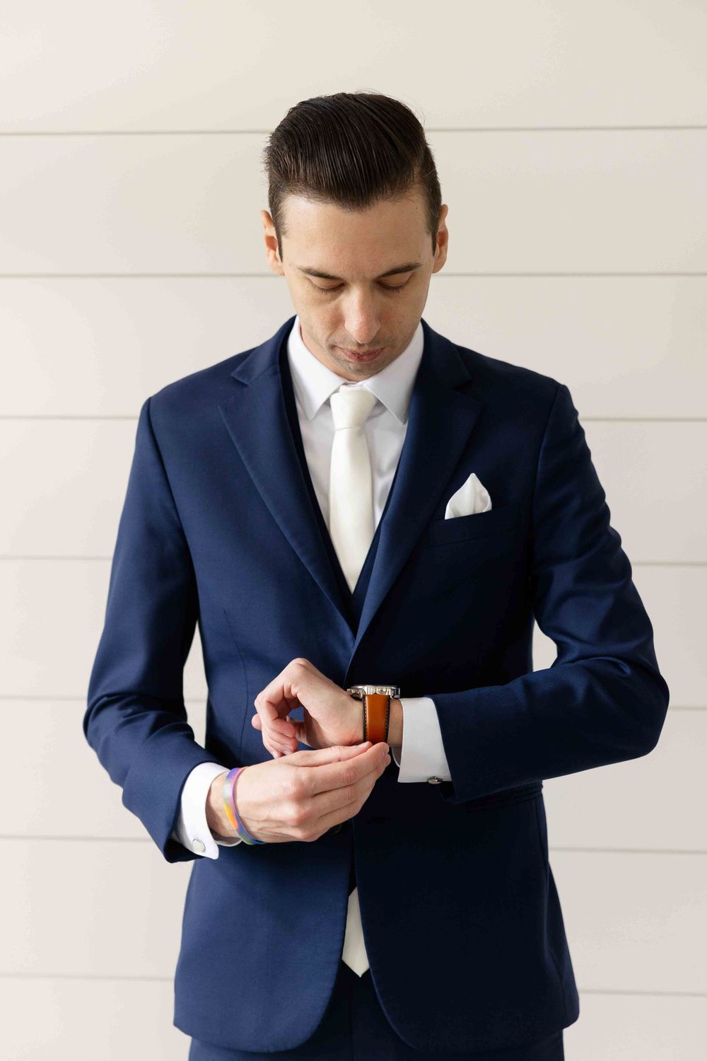 A groom in a tailored navy blue suit and white tie checks his watch against a light-colored wall.