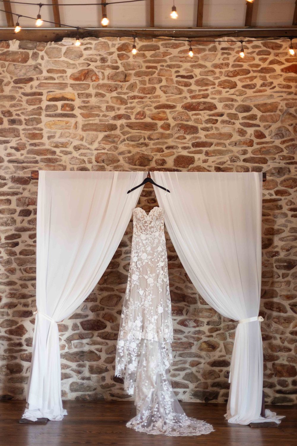 A delicate lace wedding gown displayed on a hanger against a rustic stone wall with white draping fabric panels.
