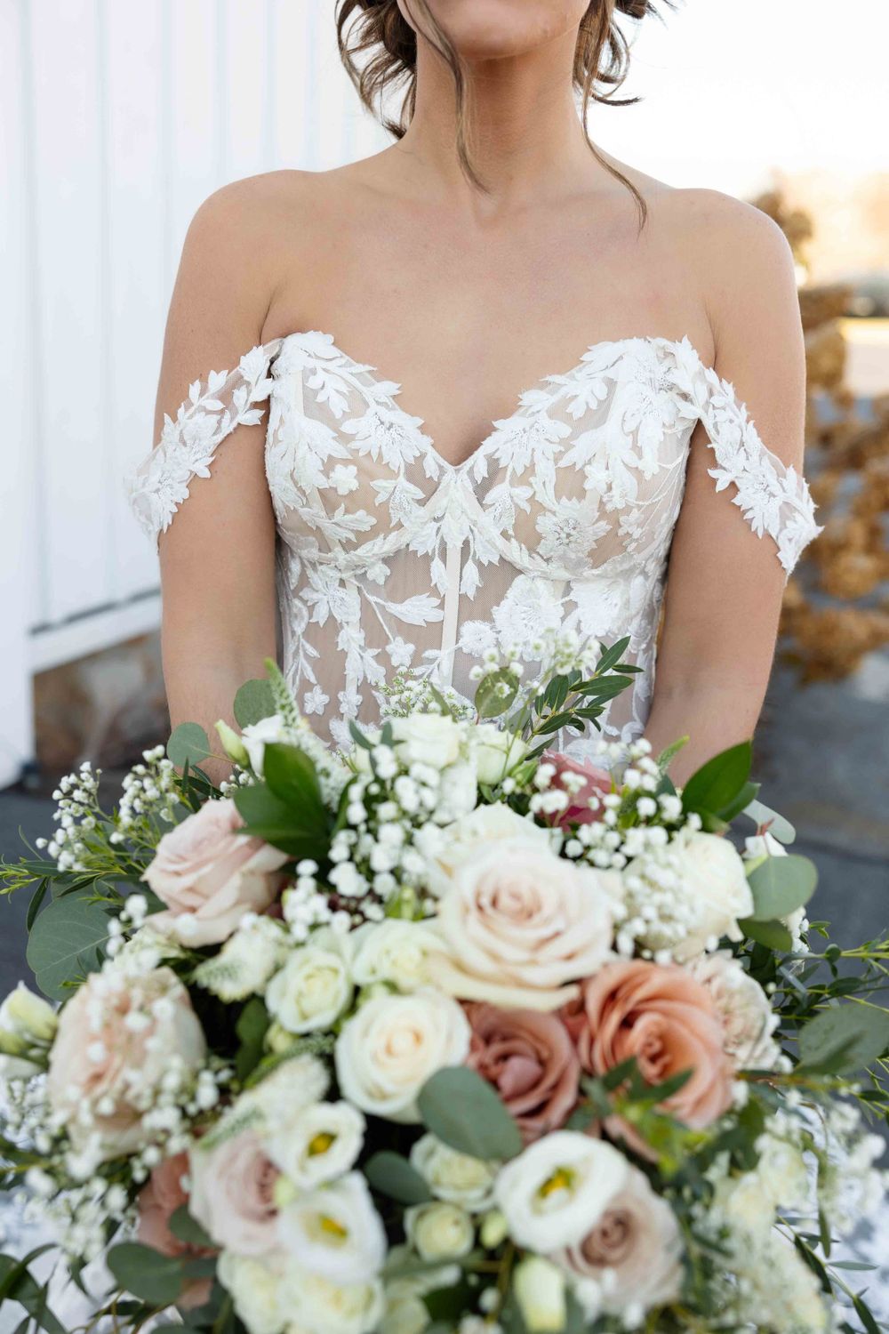Close-up view of a bride holding a lush bouquet with blush roses, white baby's breath and greenery in front of a lace bodice.