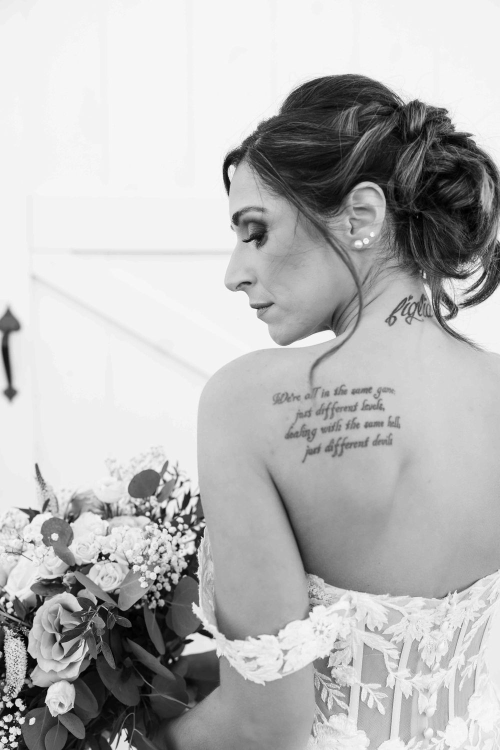 Black and white portrait showing backside of bride with script tattoo and white lace dress holding floral bouquet.