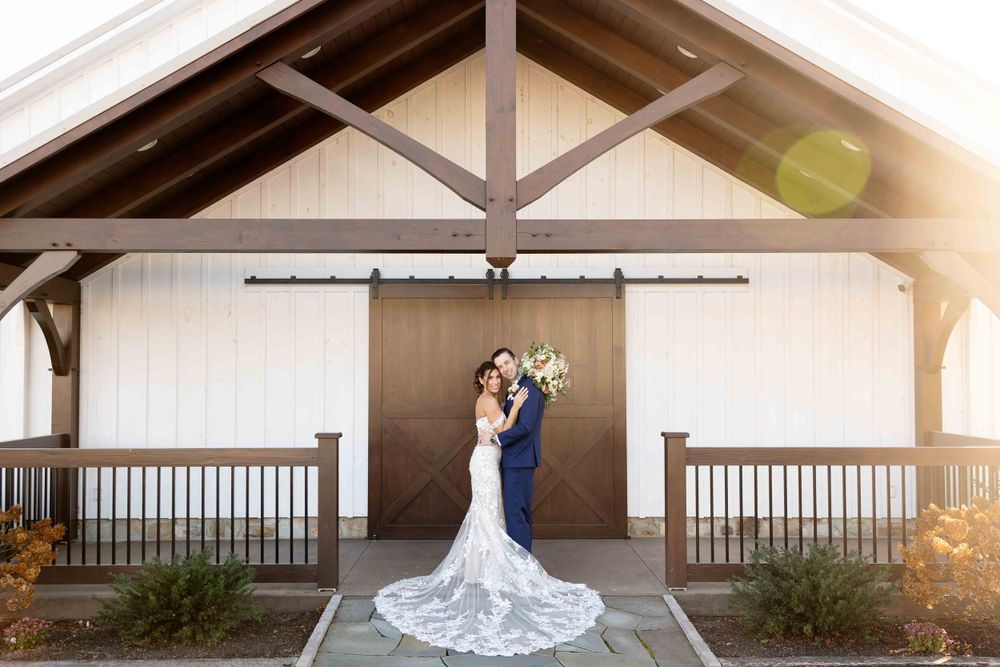 Newlywed couple posing under rustic wooden beams with sun flare and a flowing white wedding dress train.