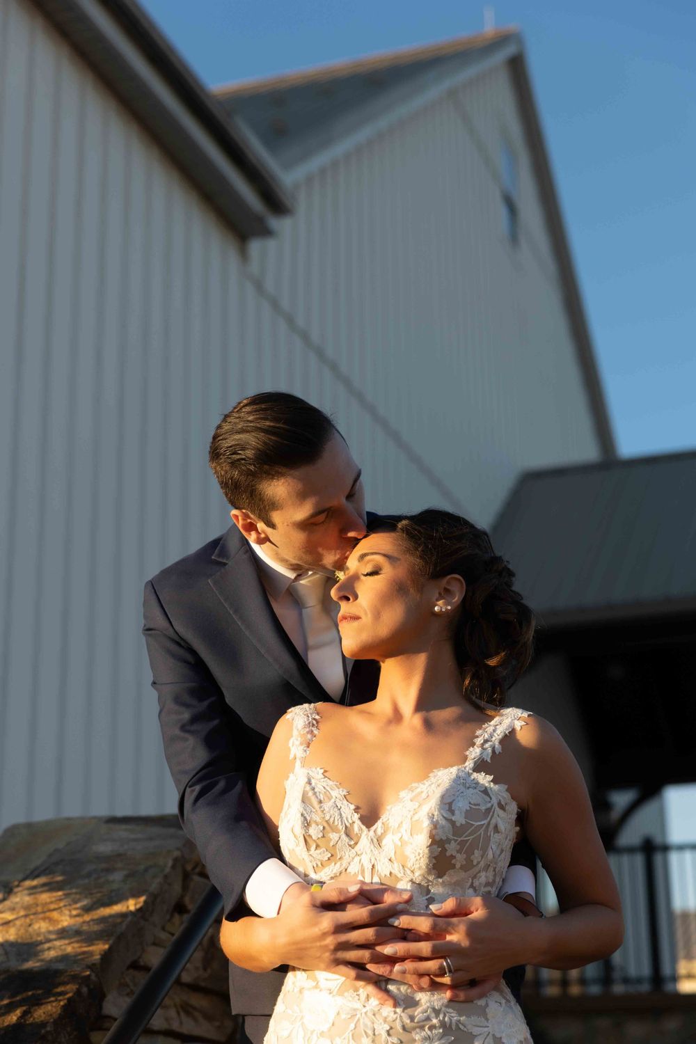 A romantic wedding couple embraces in front of a white barn during golden hour lighting.