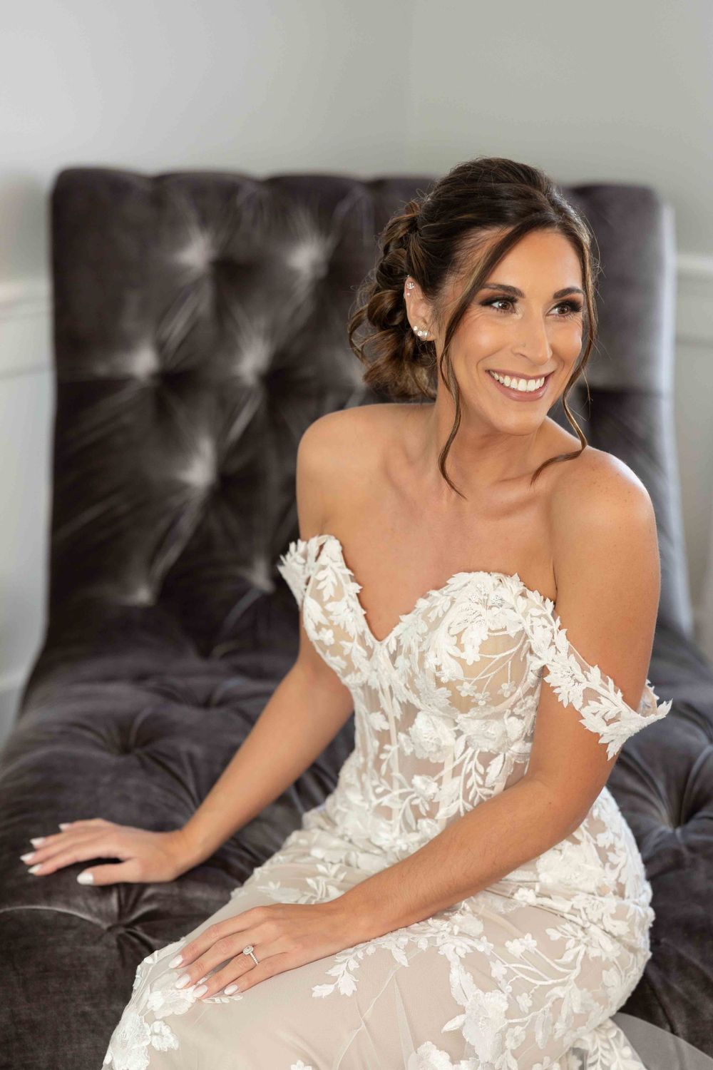 Beautiful bride wearing an ornate off-shoulder lace wedding gown sits on a dark tufted couch while smiling.