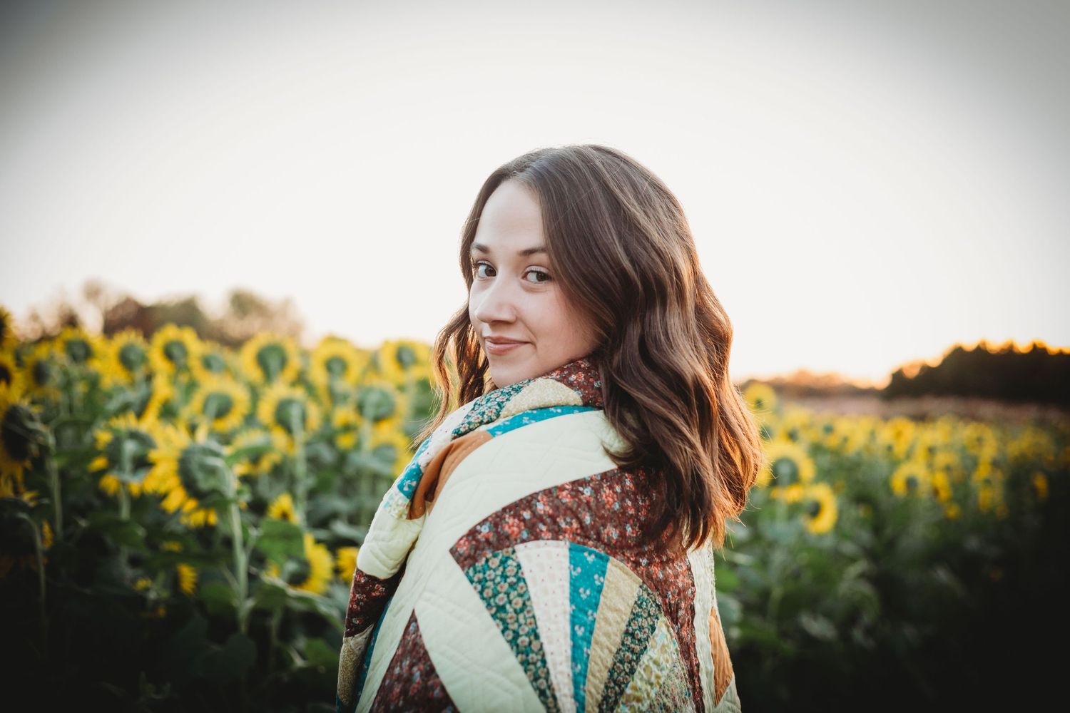 Young woman in a cozy blanket stands beside a vibrant sunflower field at sunset.
