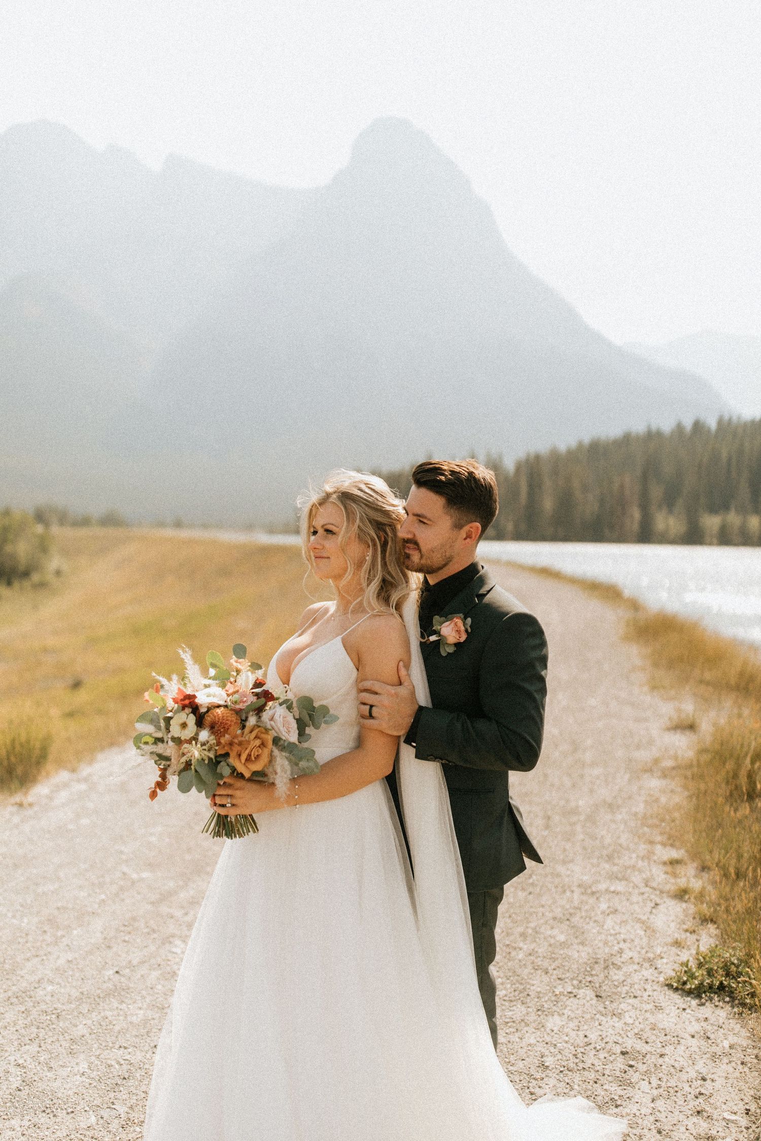A couple poses on a mountain path with a stunning alpine lake and peaks in the background.