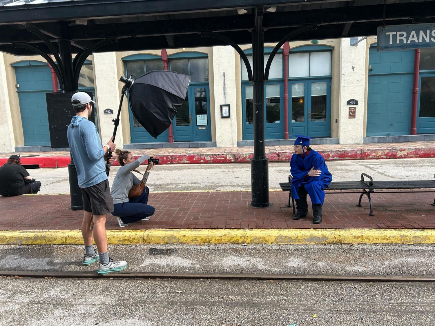 Camera crew with umbrella films interview on sidewalk outside historic storefront building during daytime.