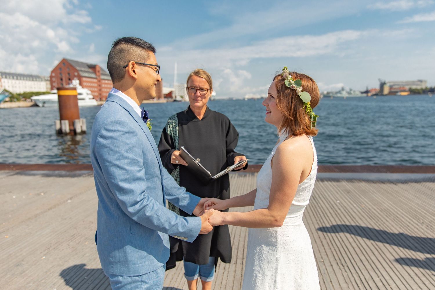 A waterfront wedding ceremony takes place on a dock with boats visible in the harbor background.