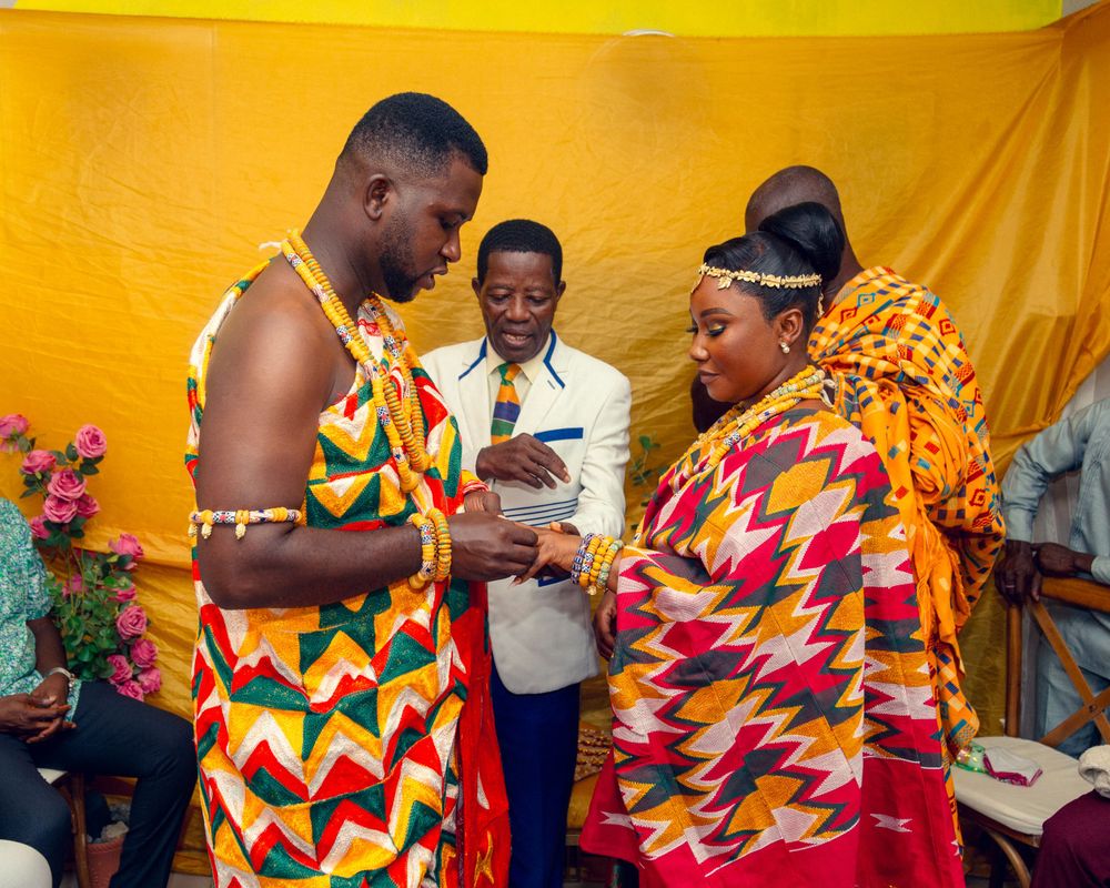 Two people wearing vibrant kente cloth patterns stand together at a gathering with a yellow backdrop.