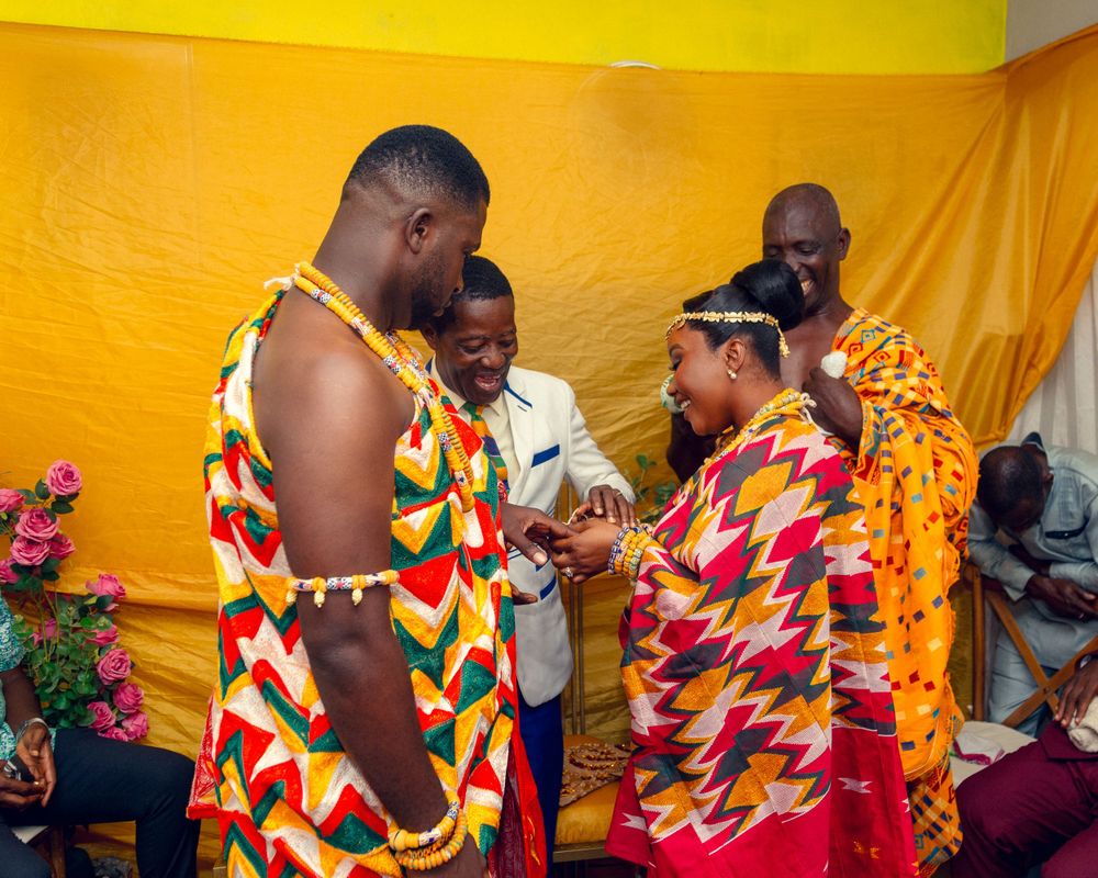 Couple in vibrant traditional kente cloth outfits participate in a cultural ceremony against yellow backdrop.