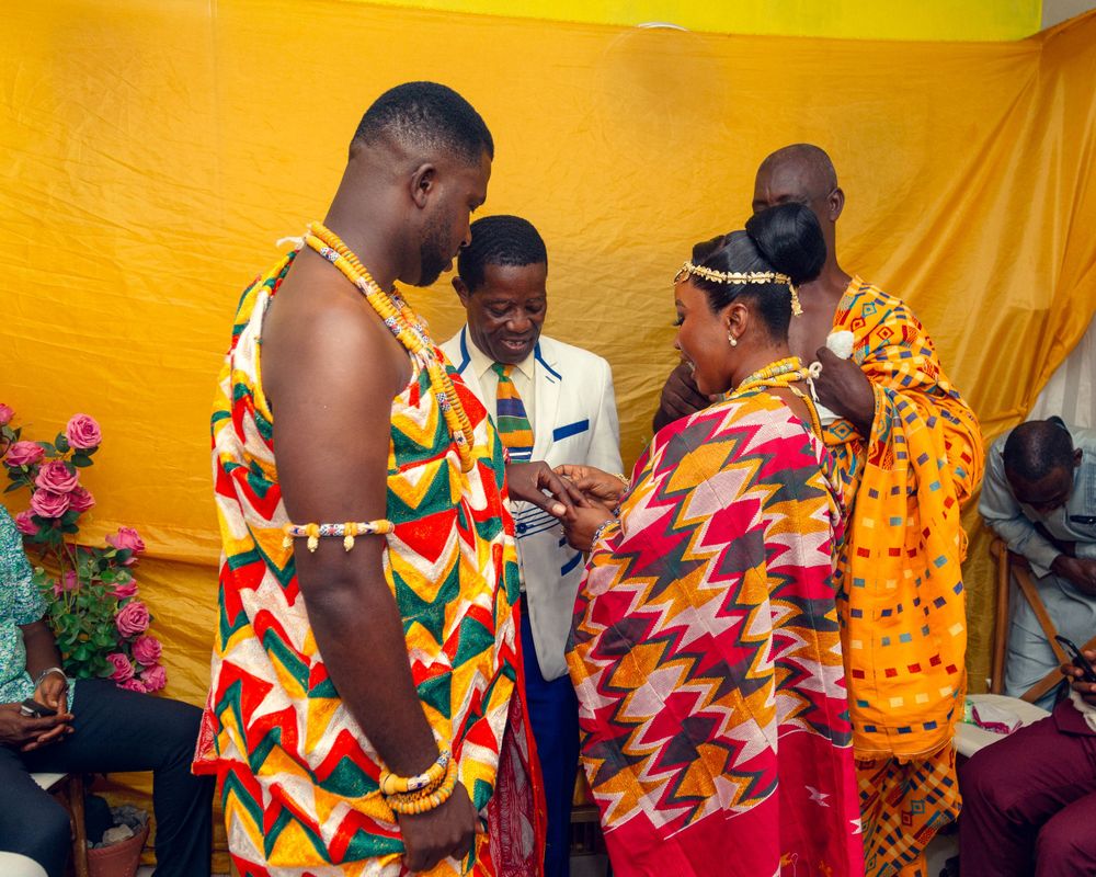 Traditional ceremony participants in colorful kente cloth share a special moment against yellow backdrop.