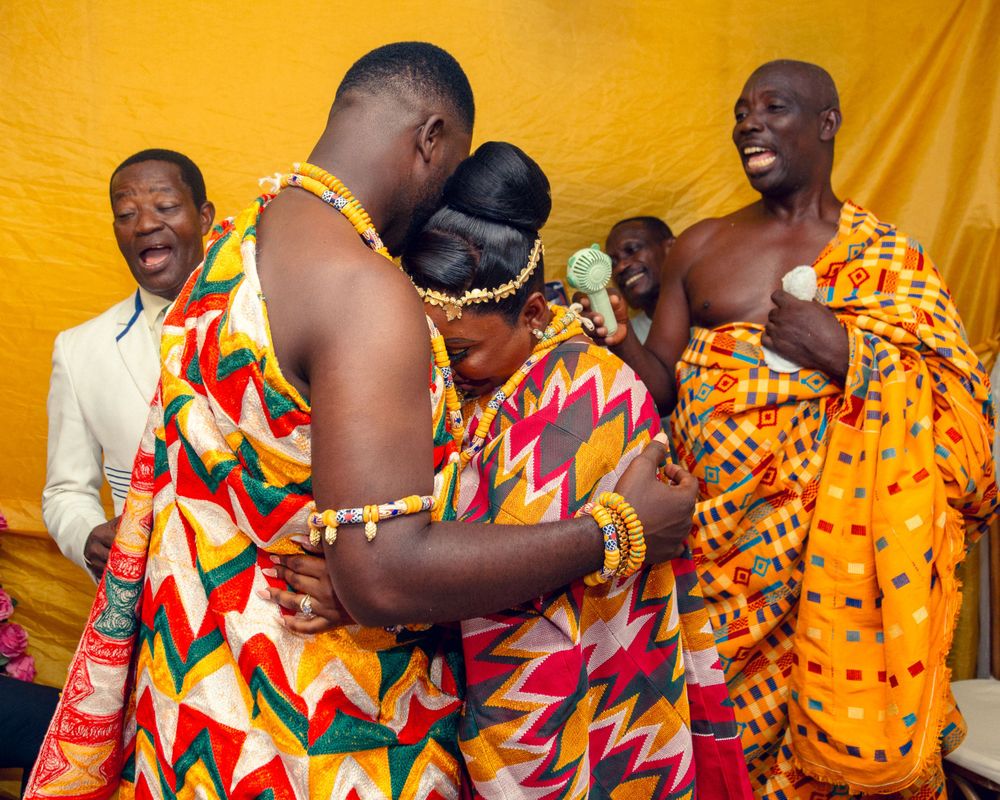 Celebratory gathering with people wearing colorful kente cloth embracing during traditional ceremony.