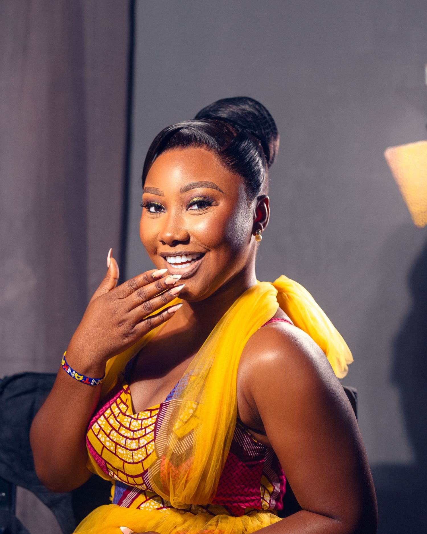 Woman in bright yellow traditional dress smiles radiantly while touching her face.