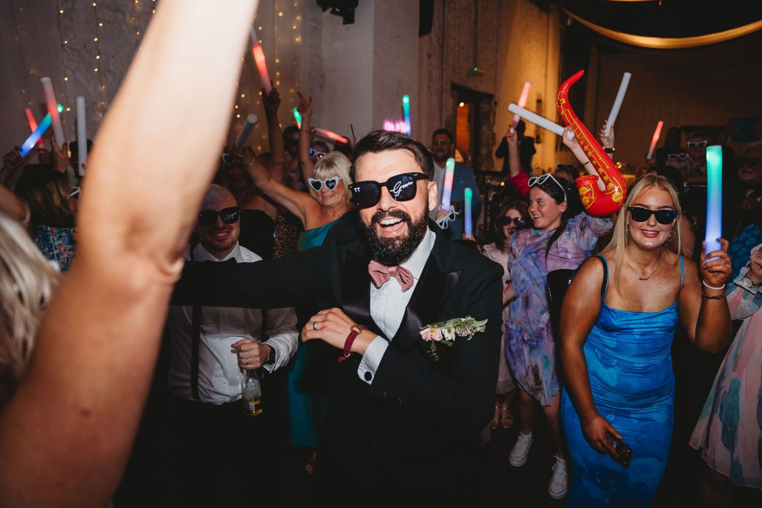 Wedding guests dance and celebrate with glowing light sticks on a dark dance floor at a reception.