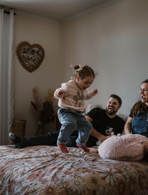 Playful toddler jumping on floral bedspread while adults look on from side of bed.