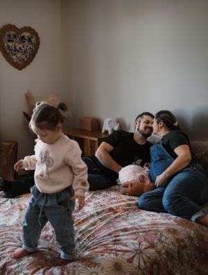 Toddler standing on floral bed while couple relaxes in background with heart decor on wall.