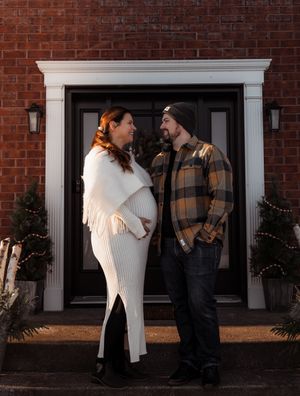 Expectant couple in winter attire standing in front of brick house with black door.